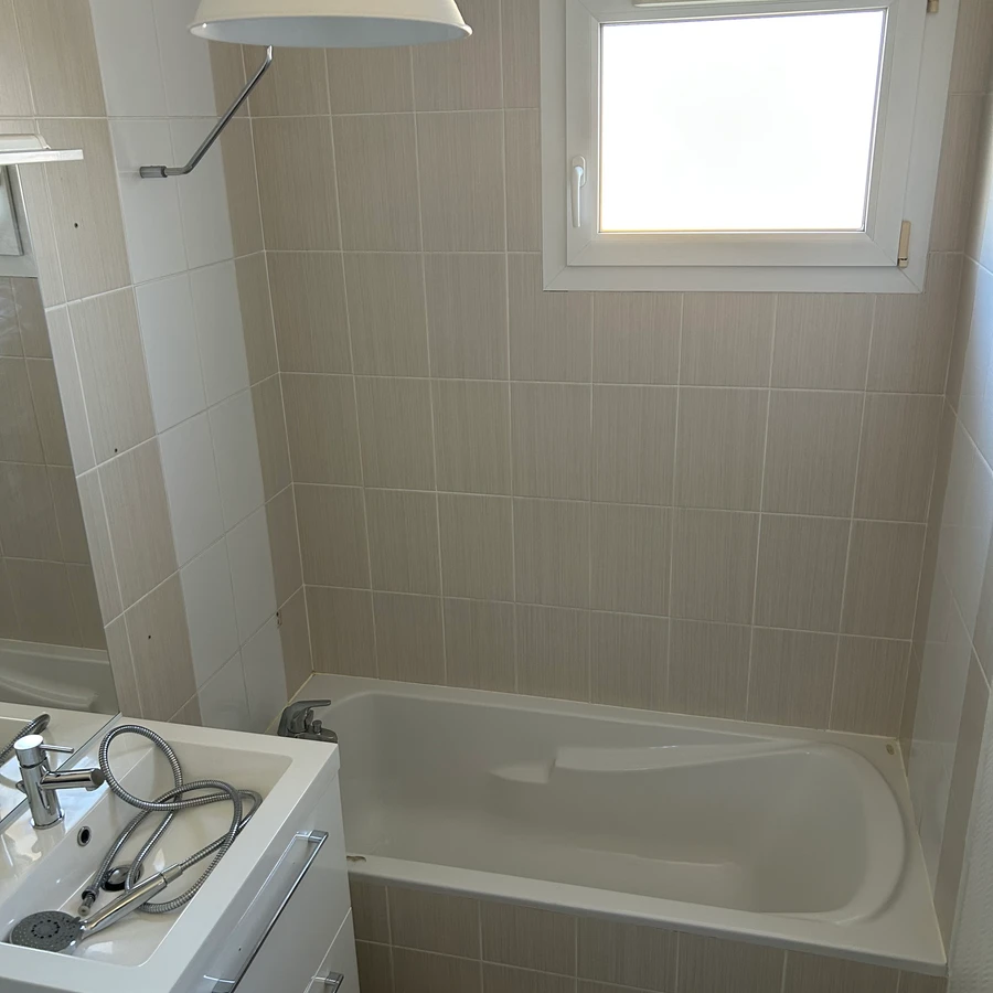 Salle de bain moderne avec baignoire blanche, carrelage beige clair, douche avec pare-baignoire et fenêtre